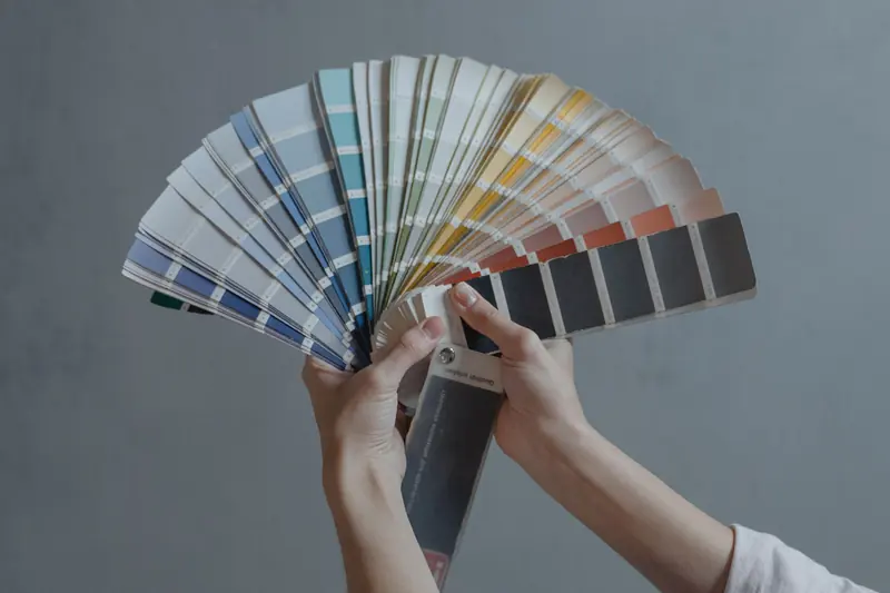 Unas manos sostienen un abanico de muestras de color frente a una pared gris.
